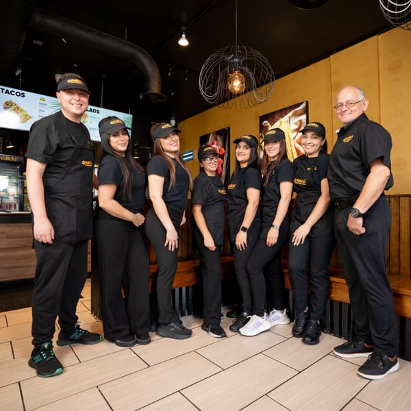 Eight MAIKKI restaurant staff members in black uniforms and visors smiling in the dining area.