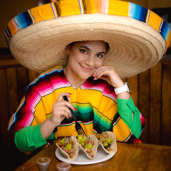 A smiling woman in a sombrero and colorful poncho sits at a table with three tacos.