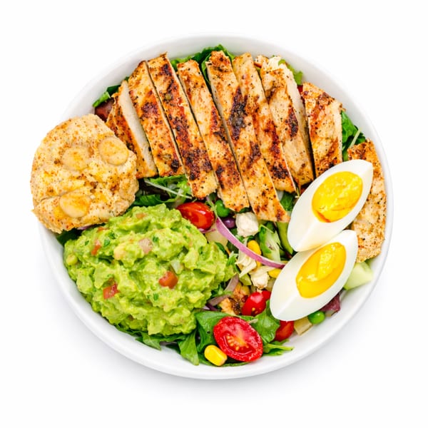 Grilled chicken salad with guacamole, sliced eggs, cherry tomatoes, and corn in a white bowl.