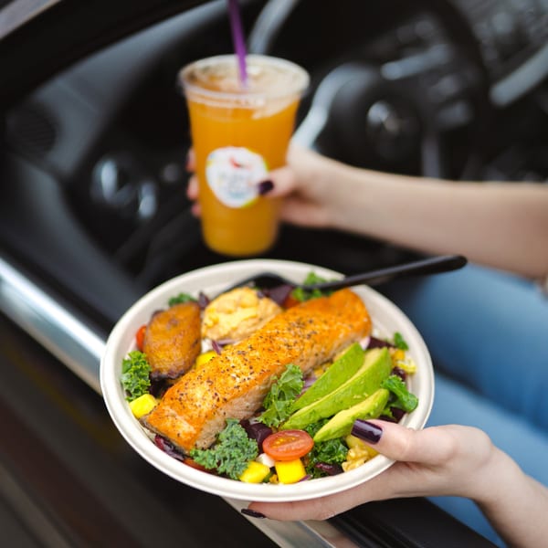 A person holds a MAIKKI salmon bowl with avocado and a tropical drink in a car.