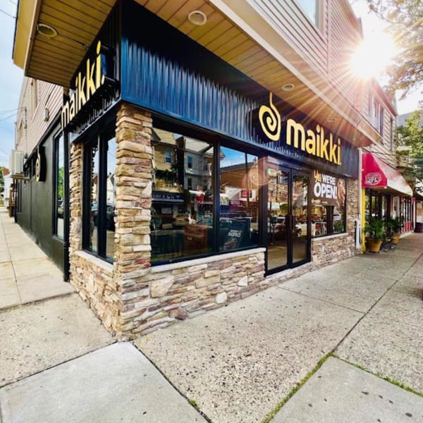 Exterior of MAIKKI restaurant with a black facade, stone accents, and large windows on a sunny day.