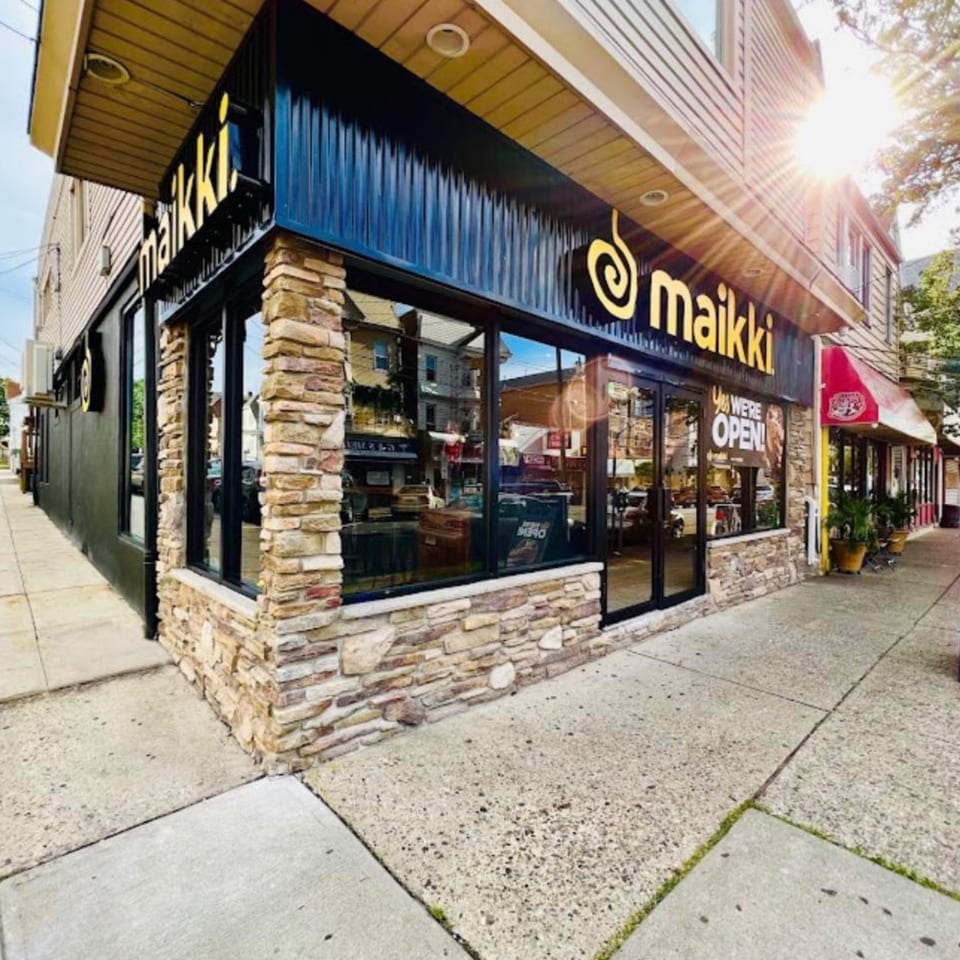 Exterior of MAIKKI restaurant with a black facade, stone accents, and large windows on a sunny day.