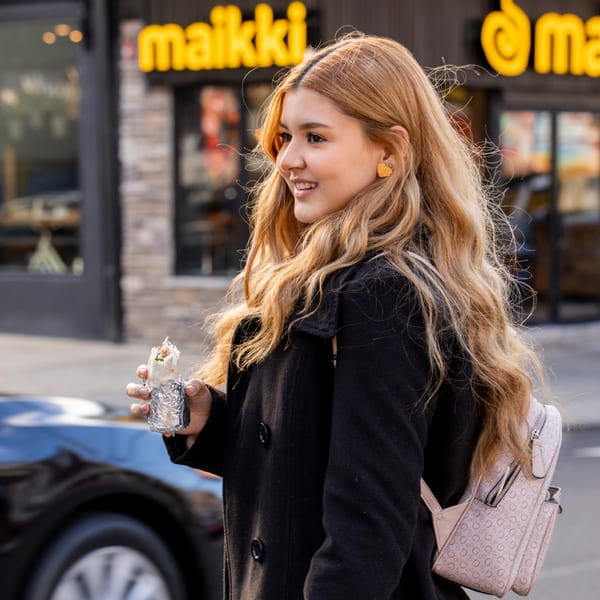 Smiling woman in black coat holding a MAIKKI burrito, with the restaurant sign in the background.