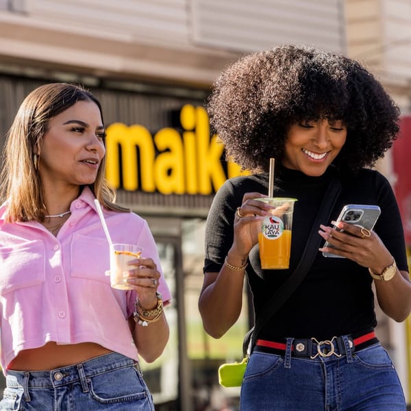 Two women holding drinks, smiling, and looking at a phone outside MAIKKI restaurant.