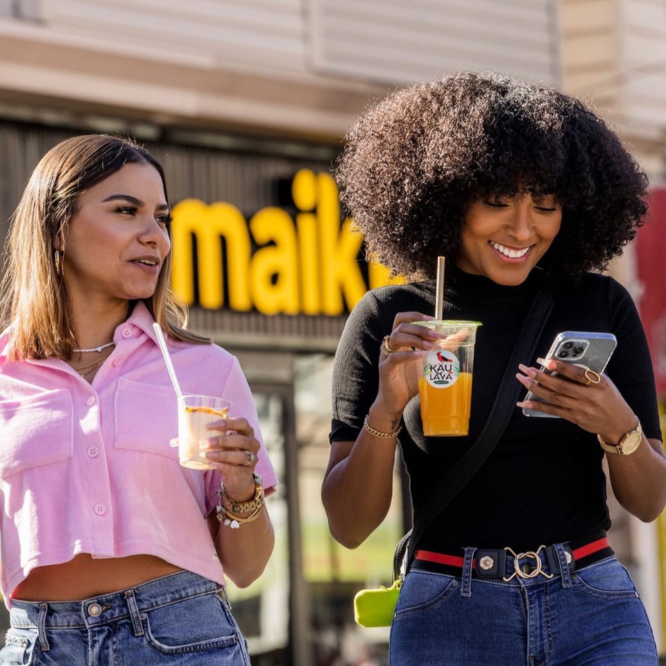 Two women holding drinks, smiling, and looking at a phone outside MAIKKI restaurant.