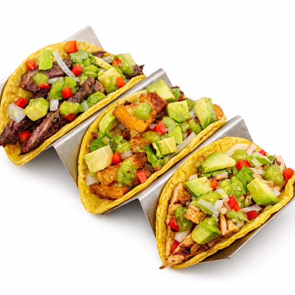 Three tacos in a metal holder, filled with meat, avocado, and salsa on a white background.